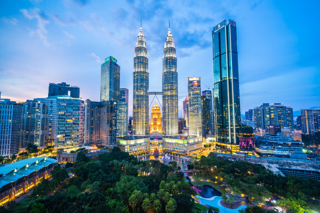 A panoramic evening view of Kuala Lumpur’s city skyline featuring the illuminated Petronas Twin Towers surrounded by modern skyscrapers and a vibrant cityscape.