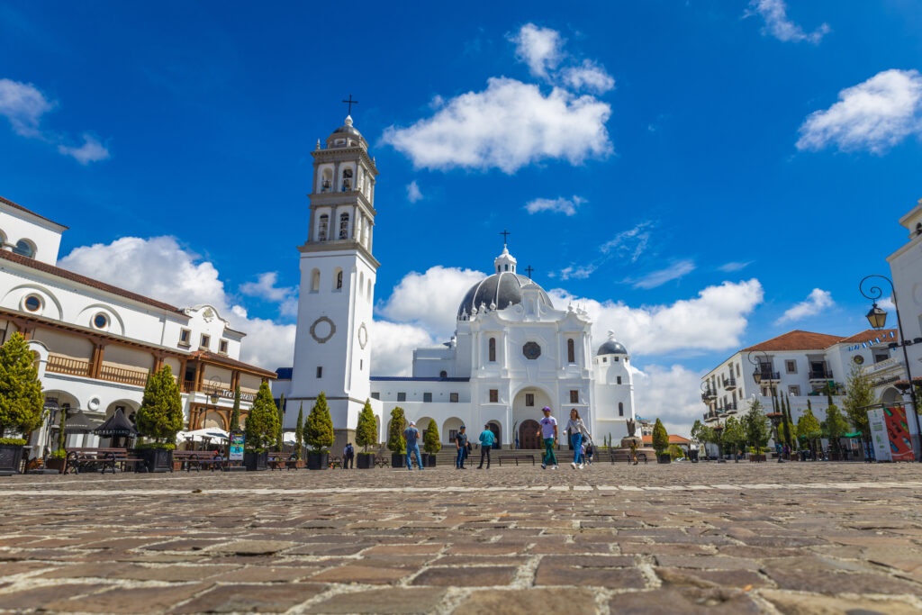 A wide-angle view of the central plaza in Ciudad Cayalá, Guatemala, featuring a white cathedral with domes and bell tower under a bright blue sky with scattered clouds.